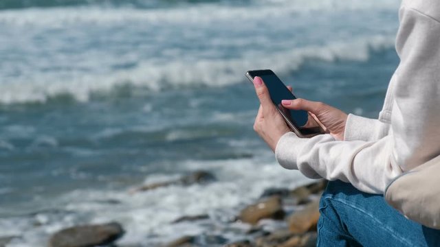 Woman is reading something on phone sitting on the sea shore in autumn. Close-up hands.
