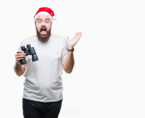 Young caucasian hipster man wearing christmas hat looking though binoculars over isolated background very happy and excited, winner expression celebrating victory screaming with big smile