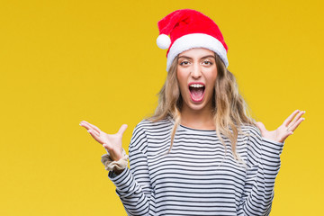 Beautiful young blonde woman wearing christmas hat over isolated background crazy and mad shouting and yelling with aggressive expression and arms raised. Frustration concept.