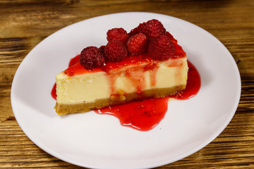 Piece of tasty New York cheesecake with raspberries and raspberry jam in a white plate on wooden table