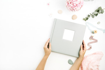 Woman hands holding wedding or family photo album. Hydrangea flower bouquet, eucalyptus branch, pastel pink blanket, monstera leaf decor on white background. Flat lay, top view still life concept.