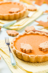 Pumpkin pie with cinnamon and cookies on yellow napkins on pastel blue background with autumn yellow leaves.