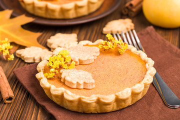 Pumpkin pie with cinnamon and cookies on brown napkins on wooden background with autumn yellow leaves.