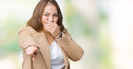 Beautiful plus size young woman wearing winter coat over isolated background Laughing of you, pointing to the camera with finger hand over mouth, shame expression