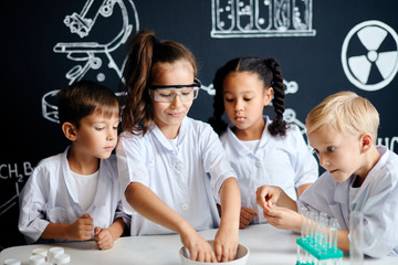 Multiethnic group of 7 years old kids in white lab coats doing a chemical experiment in laboratory...