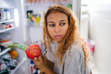 A young, tanned, sexy Asian Indonesian woman holds a fruit from her refrigerator, ready to eat for breakfast. She has blond and frizzy hair and looks calm.
