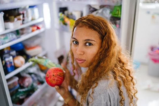 A young, tanned, sexy Asian woman holds a fruit from her refrigerator, ready to eat for breakfast. She has blond and frizzy hair and looks calm.