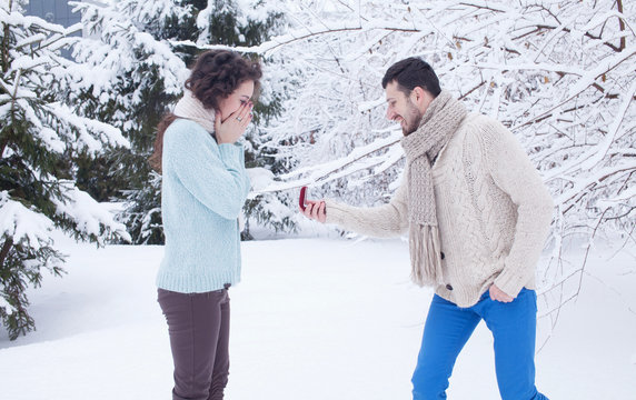 Man showing an engagement ring diamond to his amazed girlfriend in a winter park outdoor in the background. Proposal concept