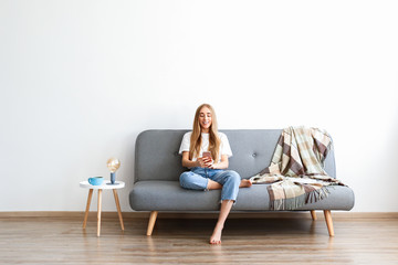 Young beautiful woman wearing white t-shirt on grey textile sofa at home. Attractive slim female in domestic situation, resting on couch in her lofty apartment. Background, copy space, close up.