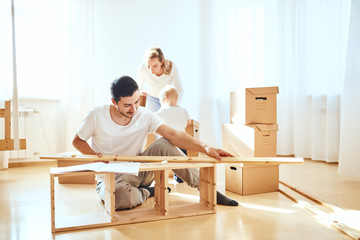 young father assembling furniture in living room of new apartment, mother with son and pile of...