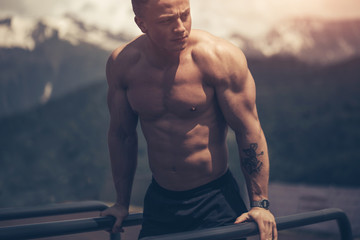 Motivated athletic shirtless man does bodyweight push ups on low horizontal bars at cool mountains...