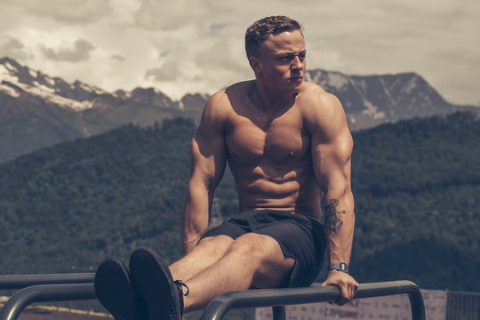 young blonde male athlete with focused and tensed face expression exercising in the park does press ups on parallel bars over mountains background .