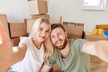 young couple is moving, making selfie, smiling while sitting among cardboard boxes