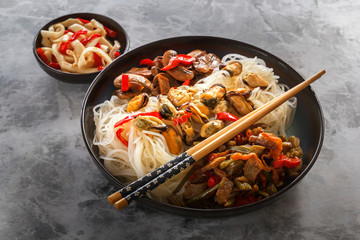Rice noodles with seafood, salad, red pepper and fried mushrooms stand on a gray table.