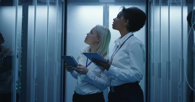 Medium shot of two women with digital tablet working in a data center with rows of server racks and checking the equipment and discussing their work