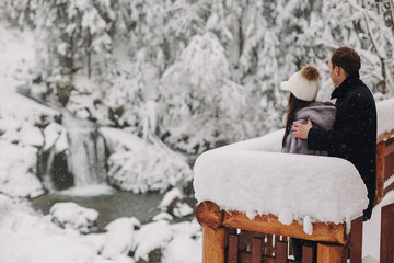 Stylish couple hugging and looking at snowy woods from wooden porch in mountains. Happy family...