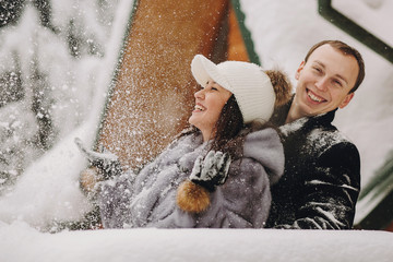 Stylish couple playing with snow in wooden cabin on background of winter snowy  mountains. Happy family having fun and smiling in snow at cottage. Holiday getaway together