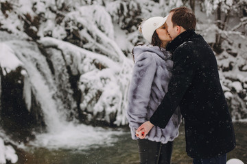 Stylish couple kissing in winter snowy mountains. Happy romantic man and woman in luxury clothes gently embracing at waterfall in snow. Holiday getaway together. Space for text
