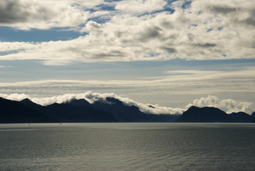 Mountains with clouds laying on mountain top under sunny blue sky with clouds looking across the ocean in Seward Alaska