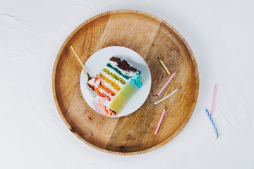 Flat lay of rainbow vegan cake piece on wooden tray with birthday candles around on grey background