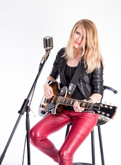 Fototapeta na wymiar blonde girl guitarist with electric guitar learns plays song, sits on chair, sings into retro microphone. Woman teacher shows how to play music. Female singer rock star in leather. rocker singer