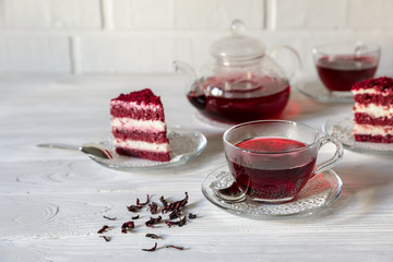 Hibiscus tea in cups and teapot, tea leaves, for dessert red velvet cake pieces on white wooden background.
