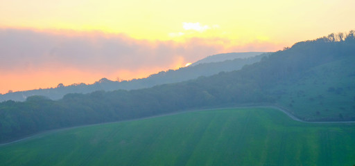 British countryside looking across a sweeping green field to a lines of trees and a hill the sun is rising behind the hill and giving an orange glow to the sky, Firle village, Sussex, England, UK,