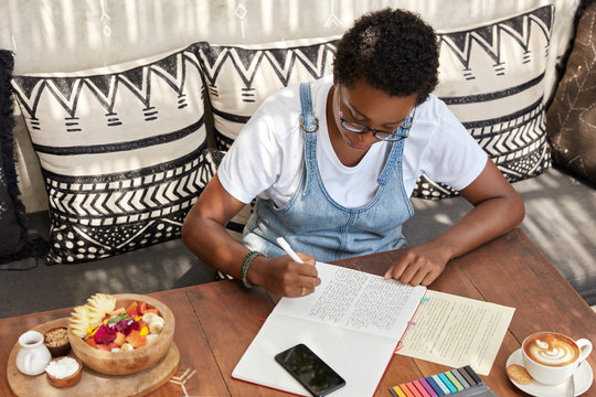Top view of attractive stylish black woman writes review of book in notepad, prepares article for local newspaper, surrounded with tasty exotic dish and aromatic coffee, enjoys comfort on sofa