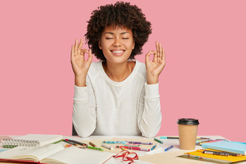 Smiling dark skinned girl enjoys tranquil atmosphere, keeps hands in meditating gesture, closes...