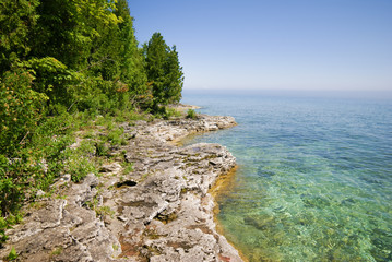 Bright blue lake to the right, gray rocky terrain with green trees on the left in Peninsula State Park in Door County Wisconsin - Powered by Adobe