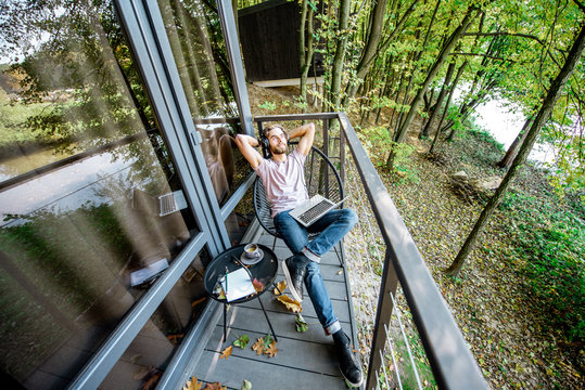 Man relaxing during the work with laptop on the balcony of his house in the beautiful forest. Wide view with reflection on the window