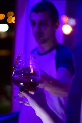 blurred young handsome man with close up  glass of red wine and female hand with wine  in the evening in the light of neon lamps
