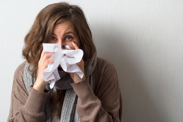 young caucasian woman with curly hair in sweater and scarf blows her nose in a paper handkerchief