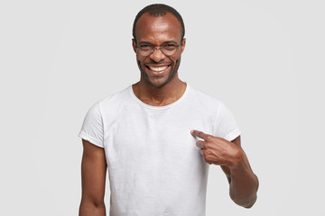 Cheerful dark skinned guy with toothy smile, points at blank space of his t shirt for your advertising content or logo, wears round spectacles, involved in advert campaign. Clothing, monochrome