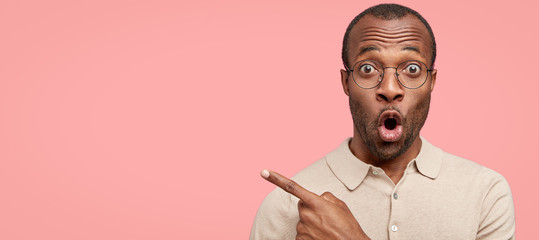 Indoor shot of emotive astonished black man opens mouth widely has bristle, wears transparent glasses, indicates with index finger aside, shows free space for your advertising content isolated on pink