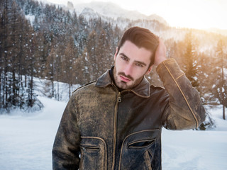 Stylish man walking confidently looking away on territory of contemporary winter resort covered in snow in the mountain, wearing brown leather jacket
