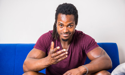 Handsome muscular black man sitting on couch at home, looking confident at camera with cute smiling expression