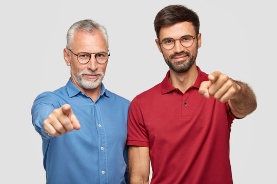 Studio shot of elderly man and bearded male adult stand next to each other indoor, point directly at camera, express their choice, indicate at something in front, have pleased facial expressions