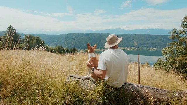 Urban nomad or adventure seeker, young man at camping spot sits on top of mountain peak in meadow overlooks alpine valley with best friend, dog partner puppy, outdoor camp vibes in nature