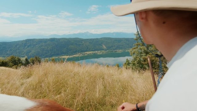 Beautiful and epic view over valley in summer with young man in outdoor camping outfit sits on log or edge in meadow or field, hugs best friend dog basenji puppy, peaceful tranquil lifestyle in nature
