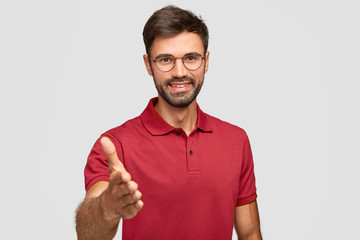 Waist up shot of happy unshaven man gives handshake, greets with someone, smiles sincerely,...