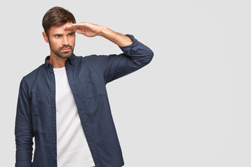 Indoor shot of unshaven man looks far, keeps hand on forehead, concentrared into distance, wears...