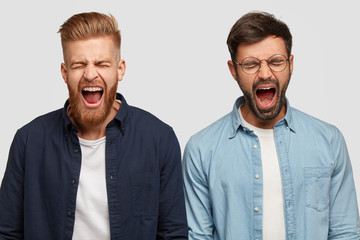 Photo of annoyed guys produce loud yell, open mouth widely, have trendy haircut and bristle, stand shoulder to shoulder against white background. People, irritation and negatie facial expressions