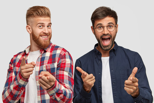Joyful males make gun gesture choose something, have toothy smile and happy expressions, dressed in fashionable outfit, model against white background, express choice. Hipsters gesture indoor