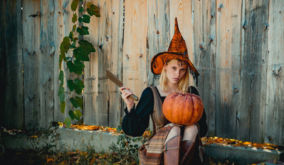 Blonde girl is preparing to carving a pumpkin. Witch posing with Pumpkin on wood background. Happy gothic young woman in witch halloween costume. Halloween concept.
