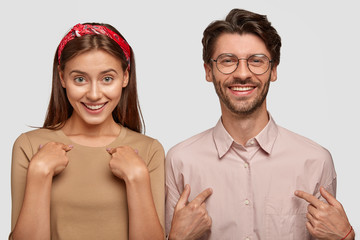 Happy smiling girl and boy have glad expressions, indicate at themselves, stand closely to each...