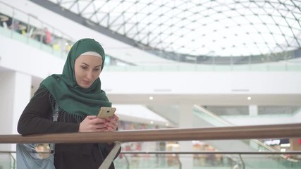 Portrait young muslim woman in a hijab stands in a shopping center and uses the phone
