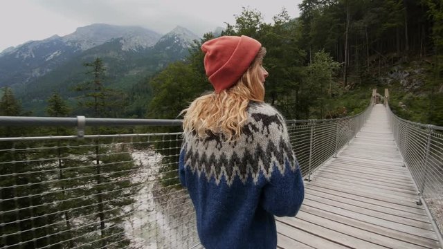 Beautiful blonde woman, female photographer in oversized wool sweater and beanie hat walks on hanging wooden bridge in mountains, in amazing and epic travel destination for lifestyle bloggers and