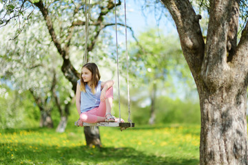 Cute little girl having fun on a swing in blossoming old apple tree garden outdoors on sunny spring day.