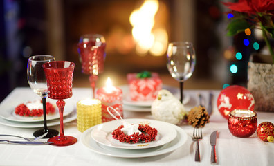Beautiful table setting for Christmas party or New Year celebration at home. Cozy room with a fireplace and Christmas tree in a background.